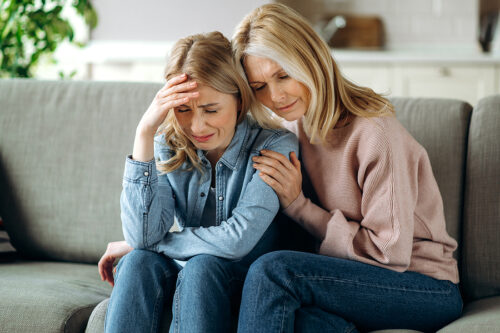 Mother and daughter together at living room. Loving mature mom sits on the couch, hugs her young adult crying daughter, stroking her, sympathizes, wrongful death claim.