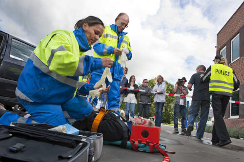 Paramedics tending to the first aid of an injured woman on a stretcher at the scene of a car crash whilst a police woman is escorting a bystander towards the cordon tape being filmed by a camera man