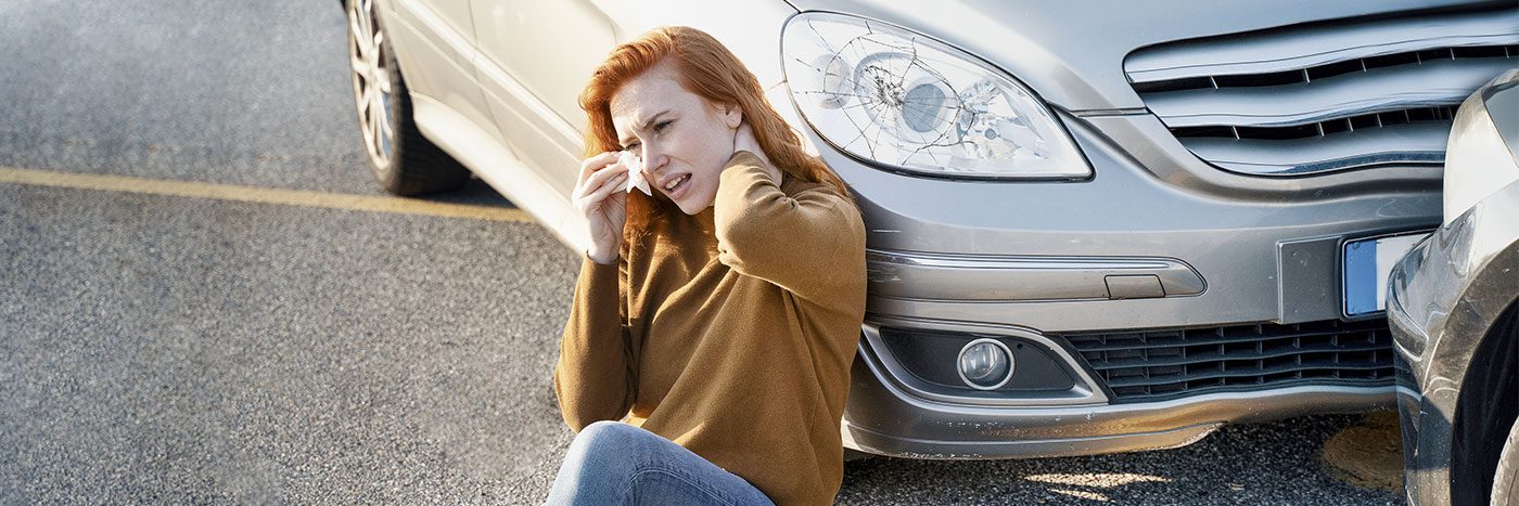 Woman who was involved in a car accident sitting by her car and holding her neck after being injured.