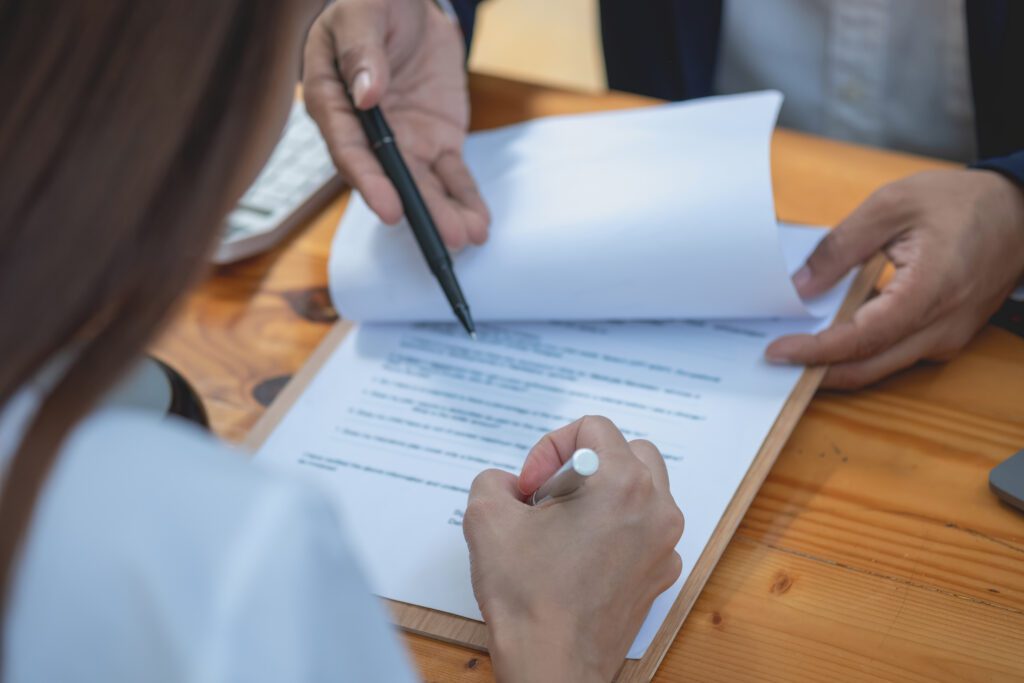 View over woman's shoulder as she signs papers for a lawyer, lawyer points at text on the paper while sitting across from the woman at the desk.