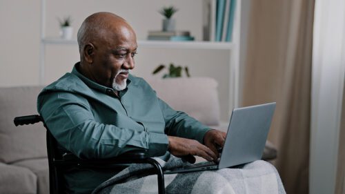 Older gentleman sitting in a wheel chair with a blanket over his legs. He is working on a laptop.