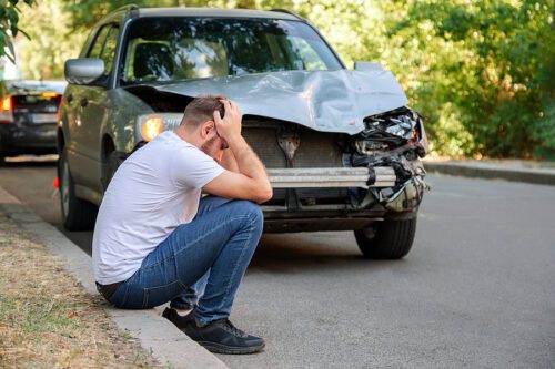 man sitting on curb holding his hands sitting in front of his damaged car after an accident