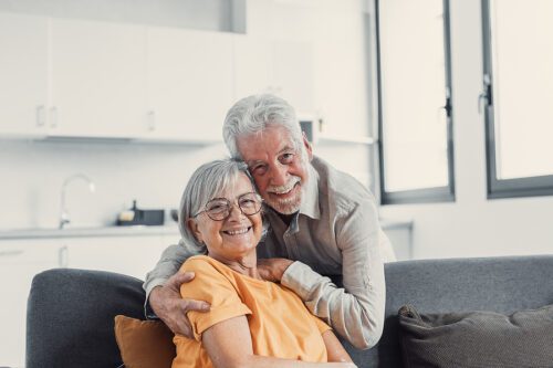 Headshot portrait of smiling elderly 60s husband and wife sit relax on couch hugging cuddling, happy mature old couple rest on sofa in living room embrace look at camera show love and care