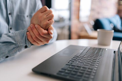 Closeup of unrecognizable mature older businessman having wrist pain during working at laptop computer sitting at desk with coffee cup. Senior business man massaging tense hand, carpal tunnel syndrome