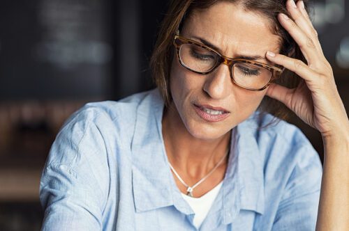 Portrait of stressed mature woman with hand on head looking down. Worried woman wearing spectacles. Tired lady having headache sitting indoors.