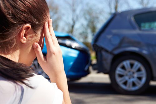 Female driver holder her hand to her forehead while looking at car accident scene and appearing stressed. 