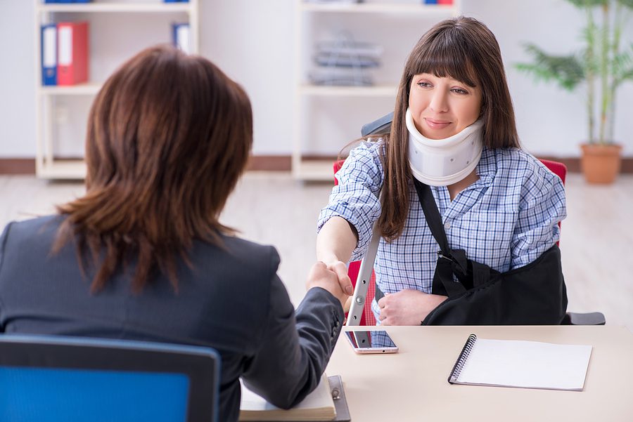 Injured woman wearing a neck brace and arm sling is meeting with a personal injury attorney and shaking her hand. 