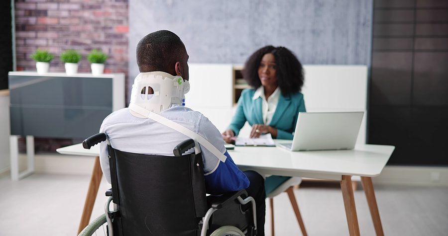 Injured man in a wheelchair wearing a neck brace, meeting with female attorney. 