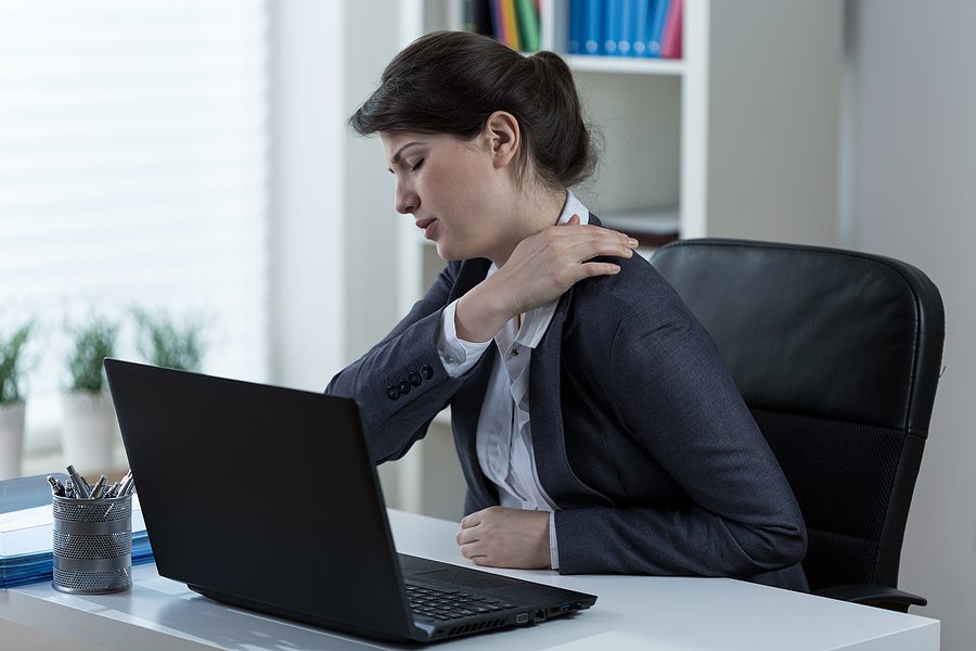 Businesswoman sitting at her desk rubbing her shoulder and appearing to be in pain. 