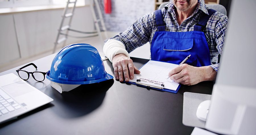 Close up of a blue hard hat on a desk with an injured male employee filling out paperwork about his injury. 