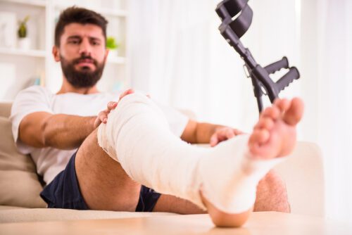Injured male sitting on couch with leg propped up and in a cast with crutches in his hand.