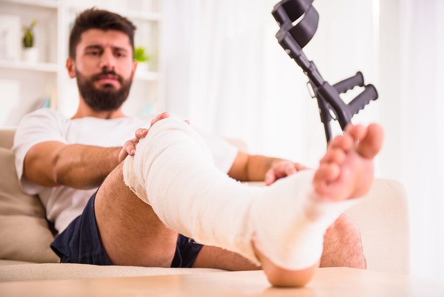 Injured male sitting on couch with leg propped up and in a cast with crutches in his hand. 