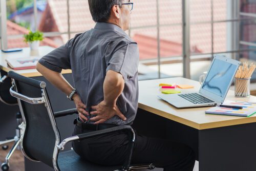 Older male businessman sitting at his desk holding his back. 