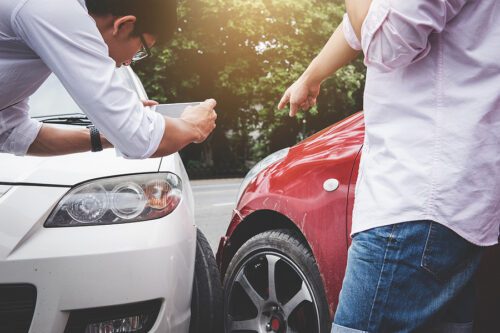 White and red sedan cars involved in an auto accident. Man takes photo of accident scene while talking with a witness. 