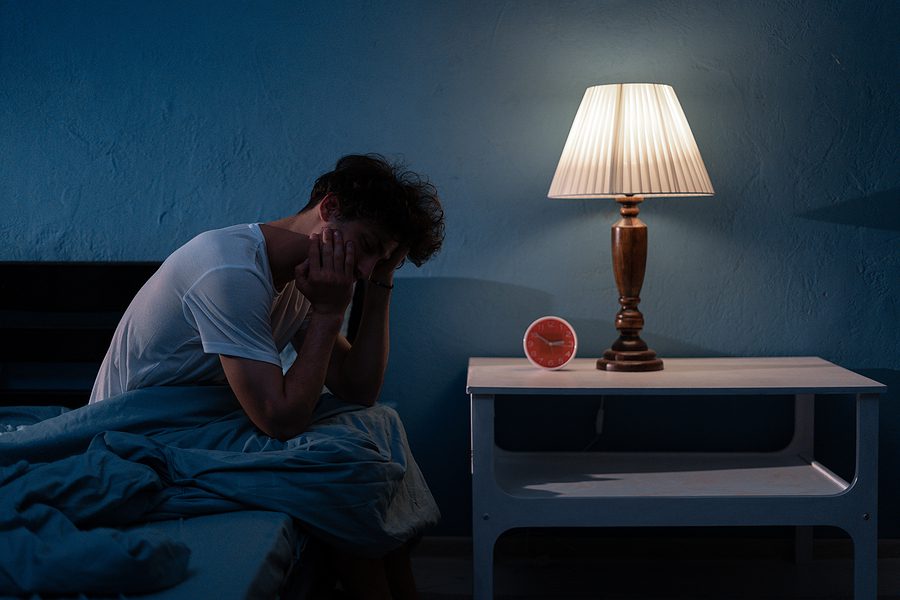 Man sitting on the edge of his bed, holding his head in his hands, not able to sleep due to stress. 