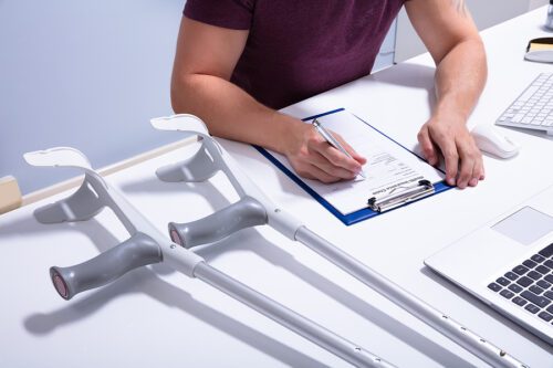 Closeup of a man filling out disability insurance paperwork with crutches lying on his desk next to him. 