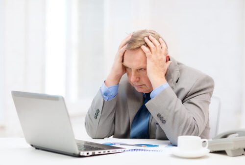 Older male sitting at his desk with his laptop open with his head in his hand and looking frustrated. 