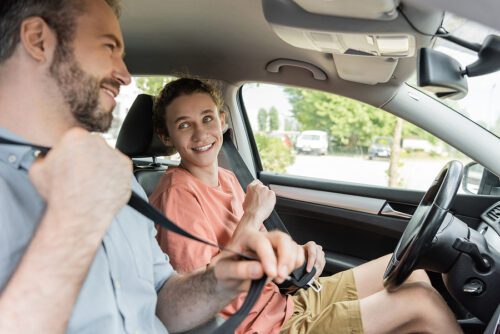 Smiling teenage boy getting ready to drive with his dad who is buckling his seatbelt. 