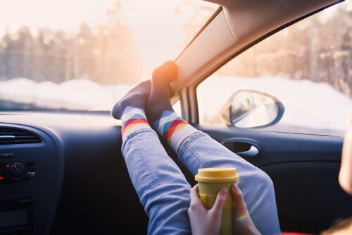 Closeup of a woman's legs and feet resting on the dashboard, while holding a cup of coffee in her hands. 