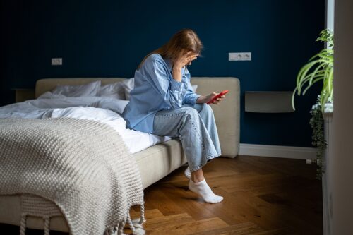 Woman sitting on the edge of her bed, reading a text message and looking stressed.
