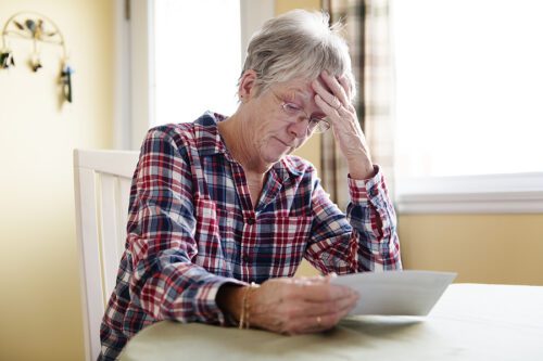 Senior woman sitting at the table reading a letter with her hand on her forehead appearing stressed. 