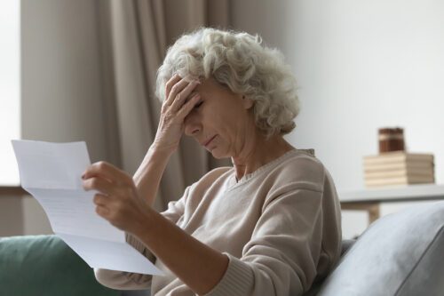 Older woman sitting on the couch, reading a letter and looking upset.